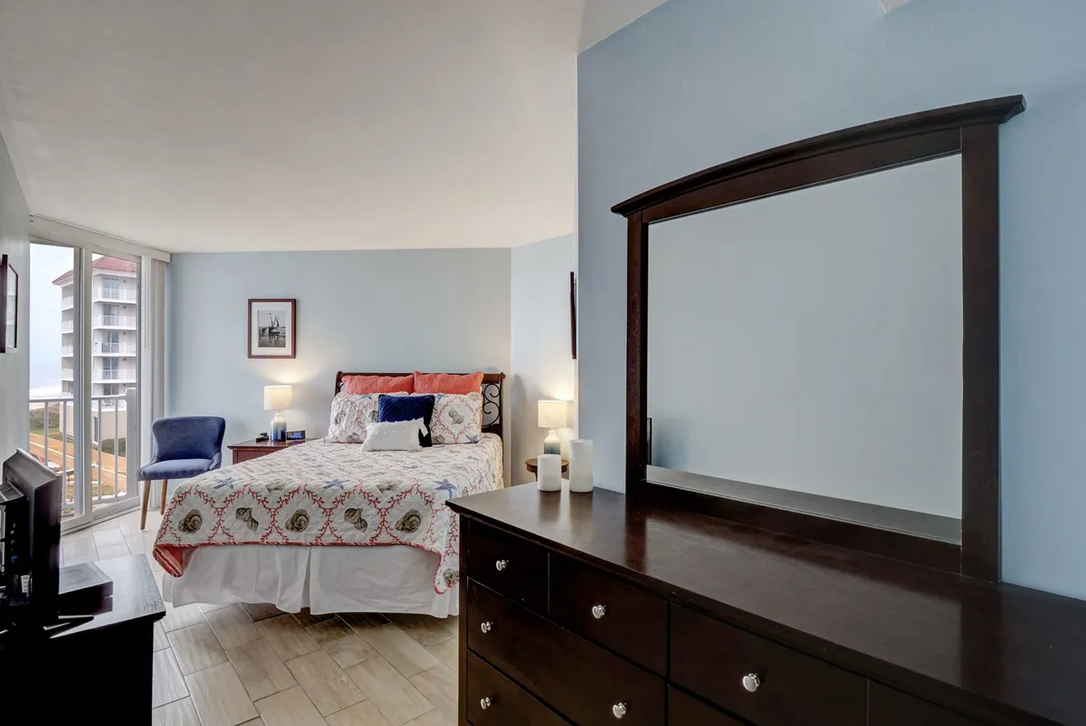 A serene bedroom featuring a queen bed with elegant, decorative bedding. Sunlight streams in through a window near an armchair, while a dressing table with a mirror and framed artwork adds a sophisticated touch. The overall décor is light, airy, and evocative of a peaceful coastal retreat.