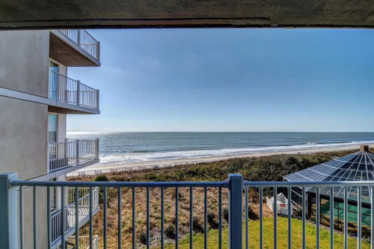 This perspective from the balcony features an unobstructed ocean view, with the sandy beach below, a sturdy railing in the foreground, and a nearby garden area. The setting captures the tranquility and beauty of the Atlantic horizon, making it an ideal spot for gatherings or quiet contemplation.