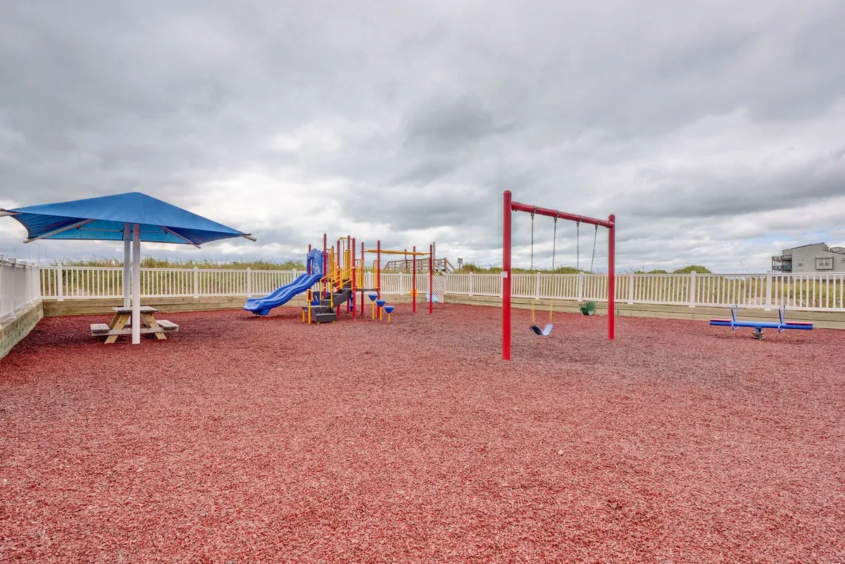 An outdoor playground featuring colorful play equipment, swings, a picnic table, and a shaded seating area. Children can be seen enjoying playtime, while adults relax and socialize in the nearby seating area.