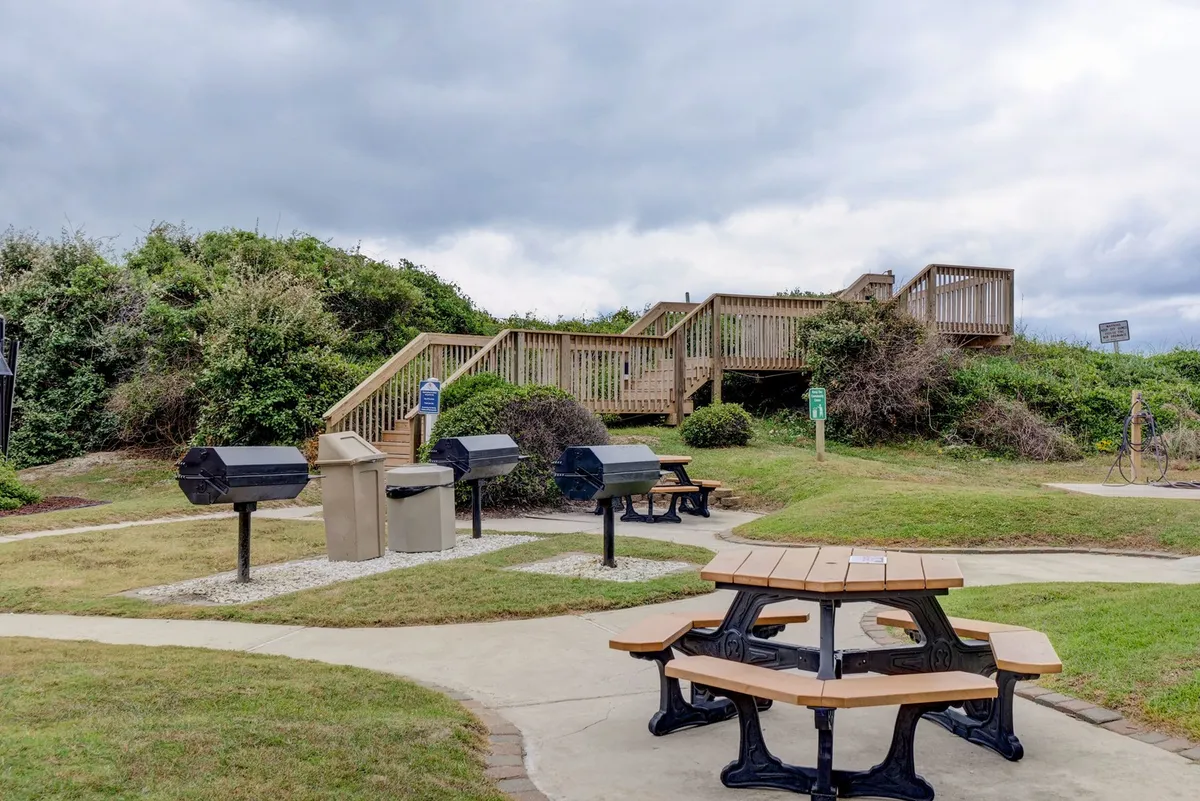 A communal outdoor area featuring picnic tables, barbecue grills, and a stairway, set against a relaxed backdrop. The space is clearly designed for group gatherings, family meals, and barbecues, with practical amenities for outdoor dining.