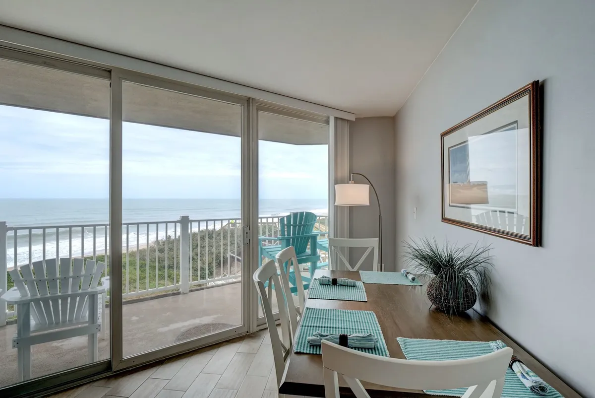 A serene dining area with a table surrounded by teal chairs, situated by large windows that offer a breathtaking ocean view. The space is filled with natural light, creating a perfect spot for meals or conversations while soaking up the coastal scenery.