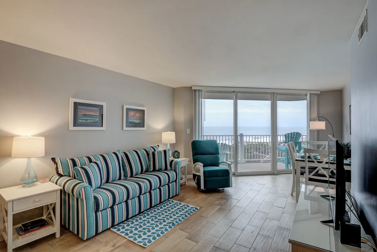 A tranquil and welcoming open-concept living and dining area, featuring a striped sofa, a dining table, soft lamps, and expansive windows that frame sweeping ocean views. The overall ambiance is light and airy, with décor that invites relaxation and togetherness.