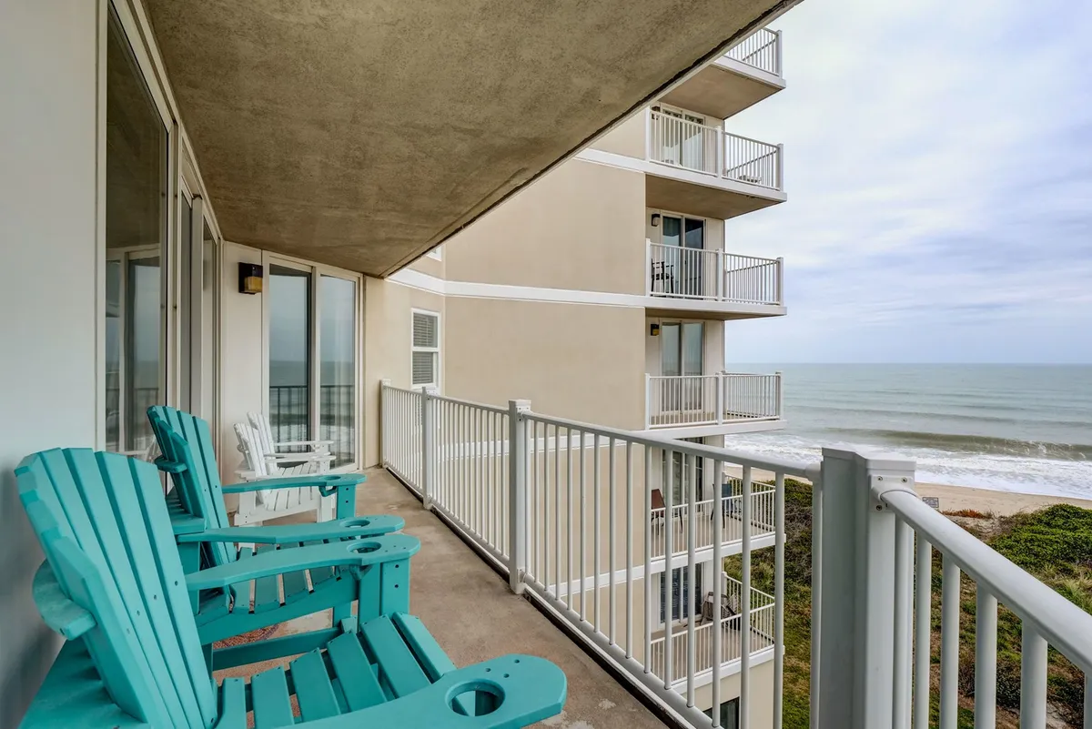 The image showcases a peaceful balcony with inviting turquoise chairs overlooking a stunning ocean view. The balcony's railing frames the endless blue waters, perfect for relaxing, reading, or savoring breathtaking sunrises and gentle evening breezes right by the beach.