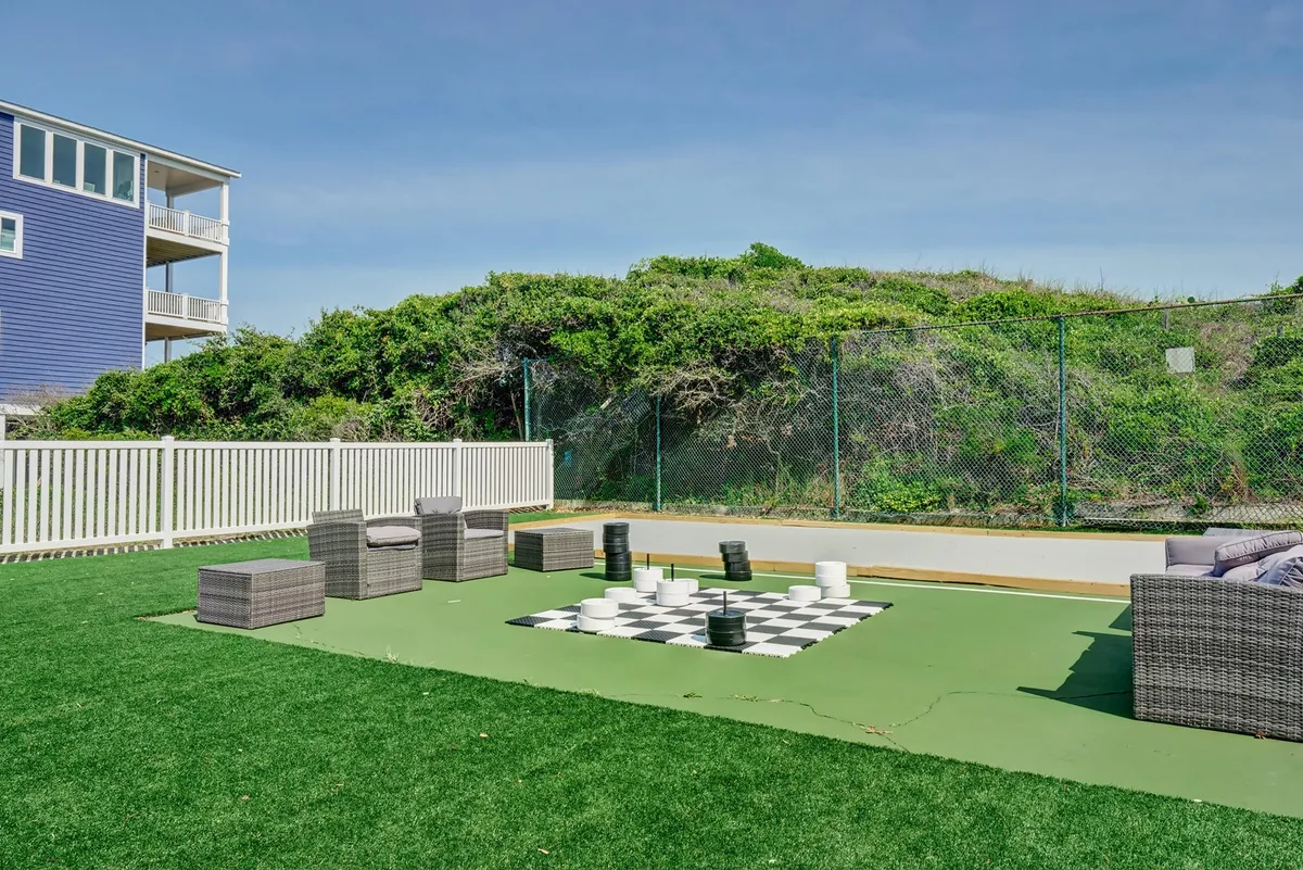 A large outdoor chess set sits on a green lawn alongside comfortable seating within a fenced area. The combination of strategic play and relaxed seating encourages both active and laid-back group interactions in a sunny, welcoming environment.