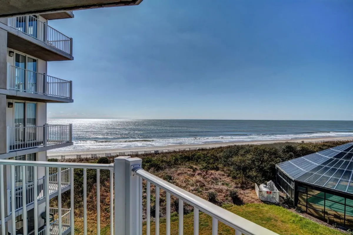 The image showcases a sweeping ocean view from the vantage point of a balcony. The foreground includes the balcony railing, while the background reveals the sandy beachfront, gentle waves, and expansive horizon. The tranquil mood is enhanced by soft lighting, inviting relaxation and reflection.