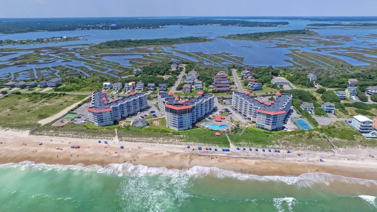 A sweeping exterior view of the St. Regis property, capturing the beachfront location, the inviting pool area, the surrounding marshlands, and the waves of the Atlantic Ocean. The lively pool scene and nearby beach make it clear how close guests are to both water amenities and nature.