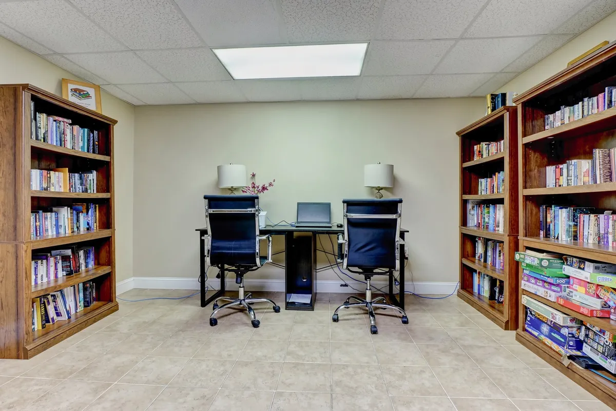 A serene, book-filled business center with two ergonomic office chairs facing a desk equipped with a computer and lamps. The environment is quiet, orderly, and bathed in natural light, offering an ideal space for productivity and remote work.