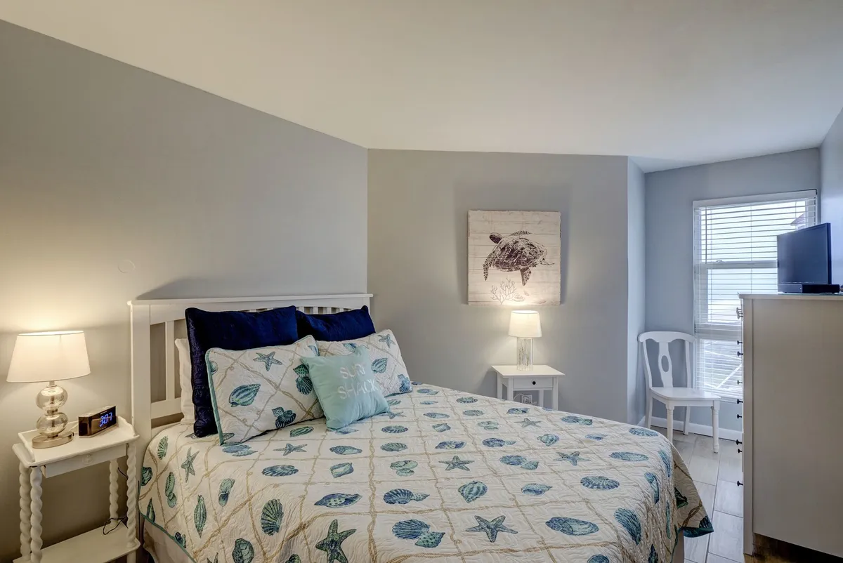 A bedroom featuring a queen bed dressed in ocean-themed bedding, flanked by side tables with lamps and decorative wall art. A small television adds to the room's comfort. The color palette is gentle and reflective of the coastal setting, offering a peaceful retreat.