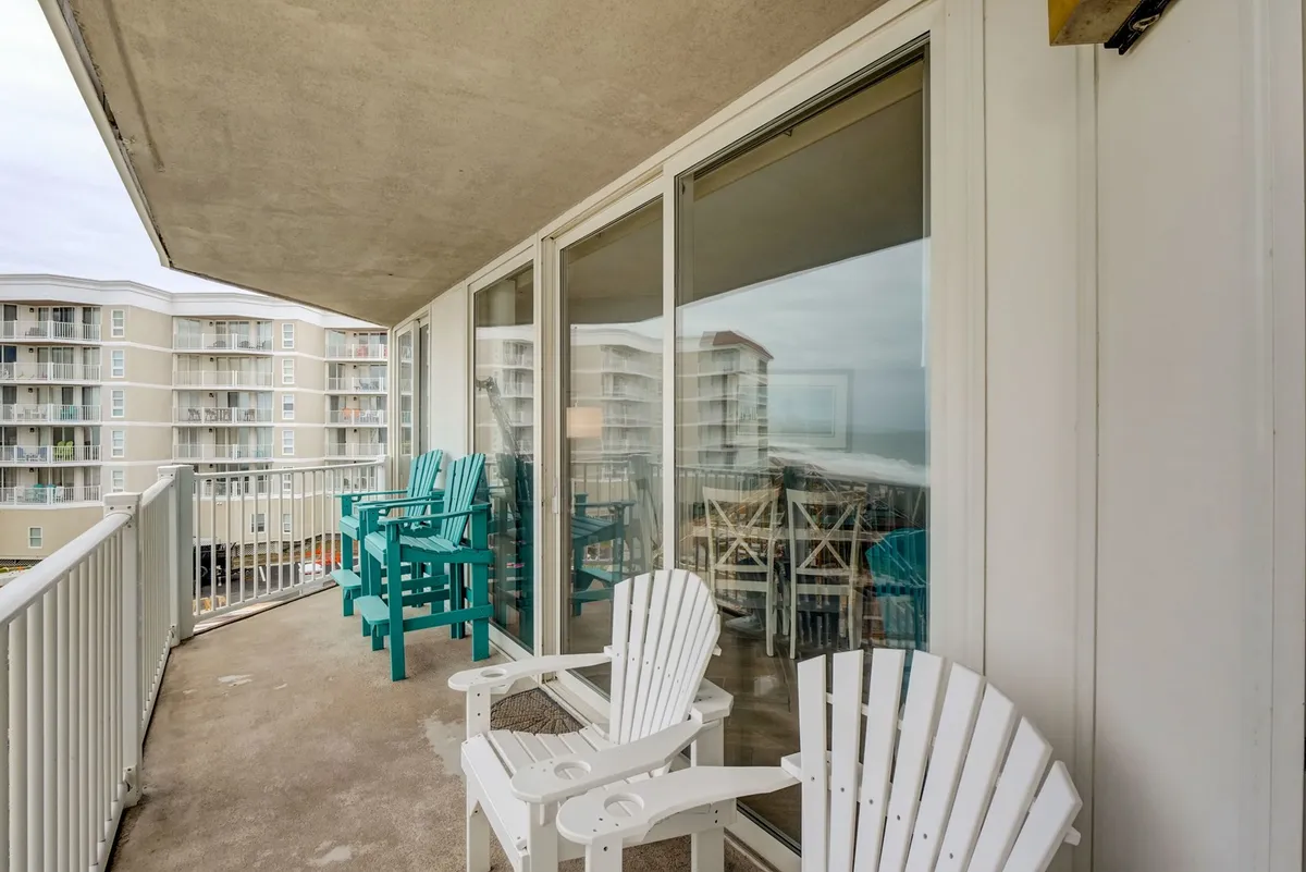 This image captures a spacious balcony outfitted with white Adirondack and teal outdoor chairs, a sturdy balcony railing, and glass doors reflecting the surrounding views. The setup suggests a perfect setting for enjoying the ocean breeze, sipping a morning coffee, or socializing with friends as the sun rises or sets.