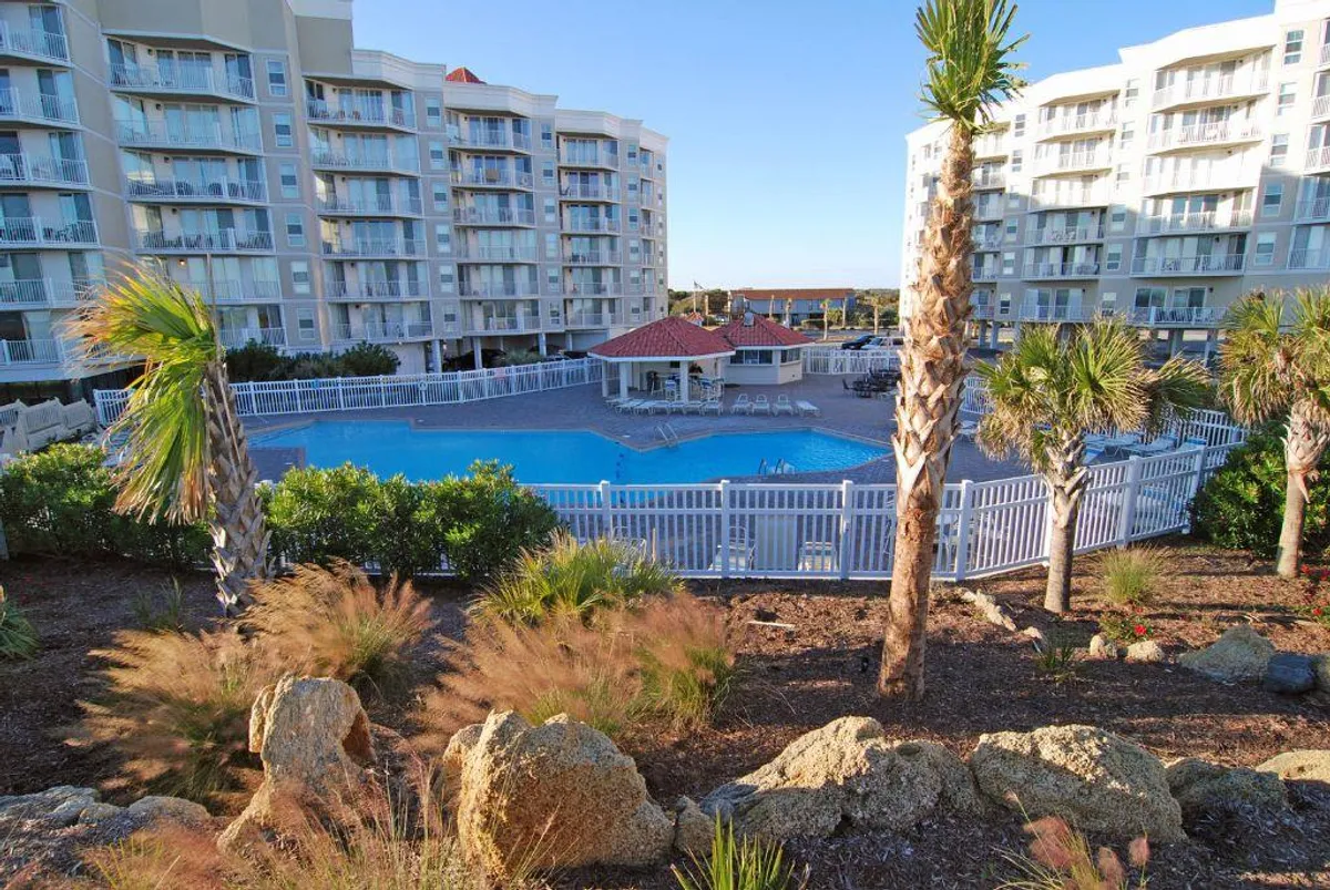 A vibrant, sunlit swimming pool surrounded by palm trees and lush landscaping, with modern resort buildings in the background. The inviting pool area features ample space for swimming, relaxing poolside, and gathering with friends and family.