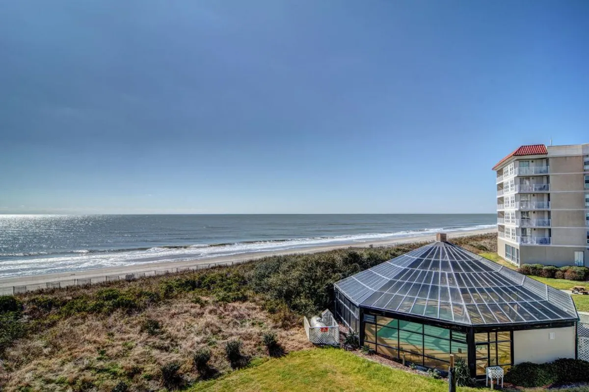 A wide, sunlit view of the Atlantic Ocean from a vantage point overlooking a glass-enclosed structure surrounded by lush greenery. The tranquil water stretches to the horizon, and the outdoor space hints at relaxation and leisure.